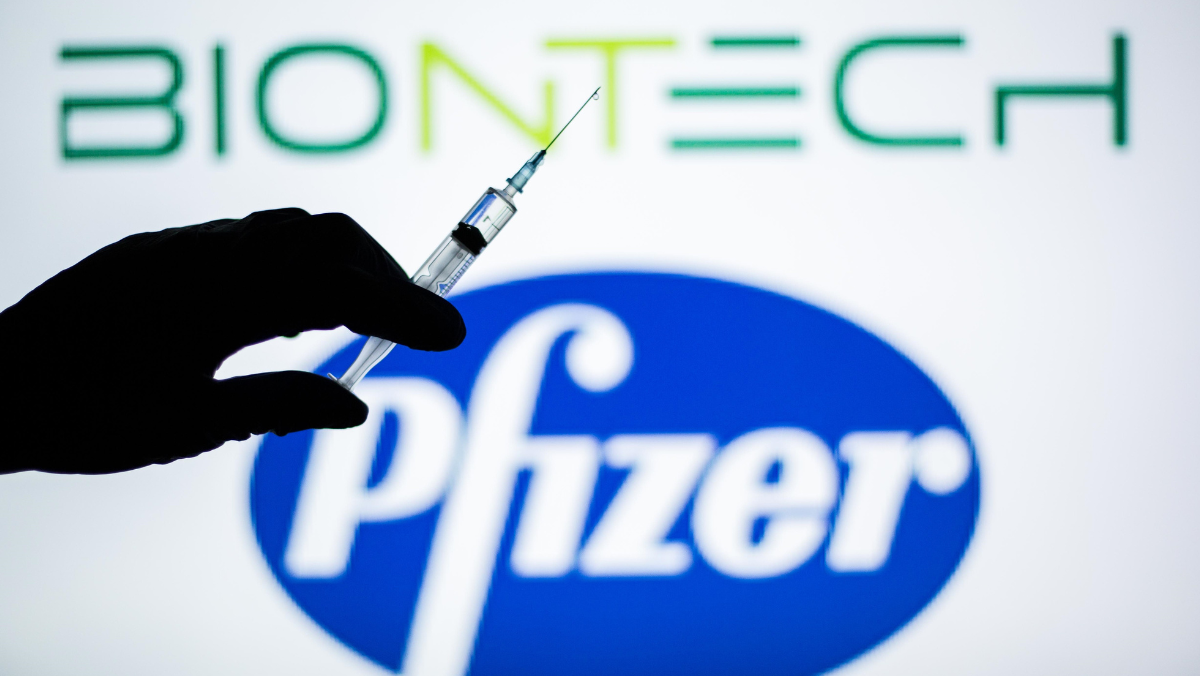 Silhouette of a hand holding a syringe in front of BioNTech and Pfizer logos on a bright display background